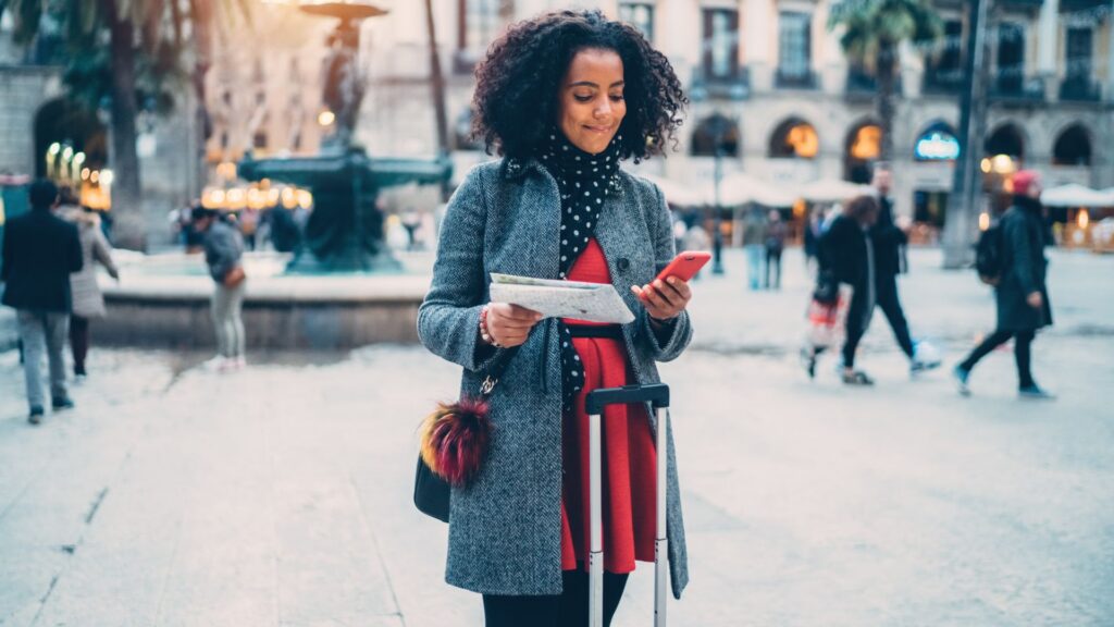 A traveler checking her phone and a paper map while pulling her suitcase through a city square overseas