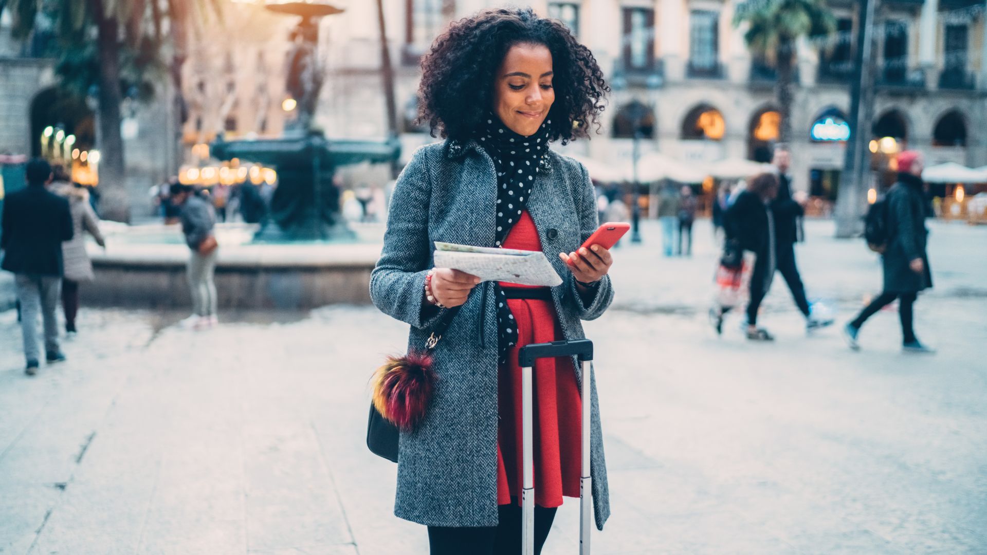 A traveler checking her phone and a paper map while pulling her suitcase through a city square overseas