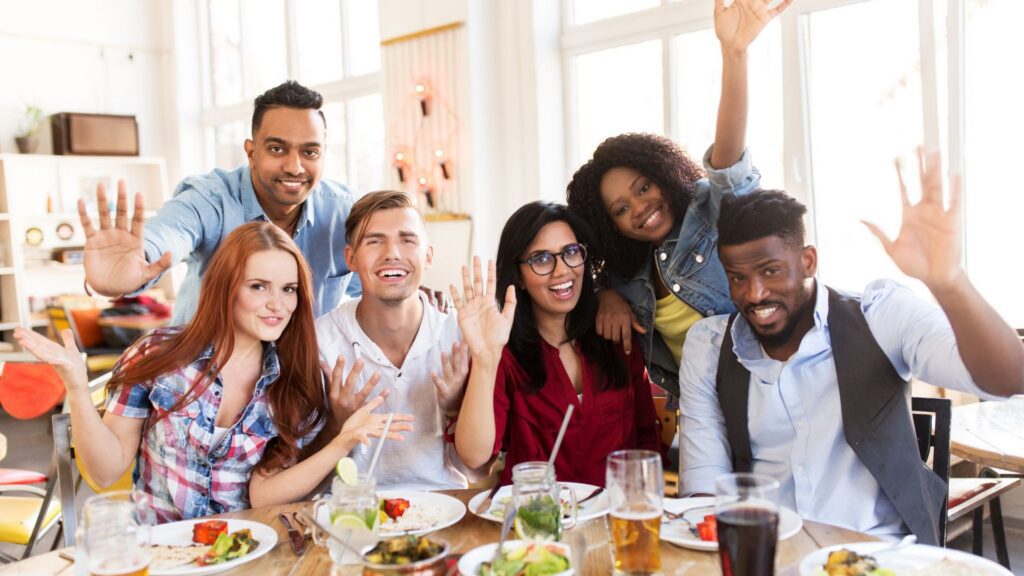 A diverse group of friends waving and celebrating together at a restaurant table.