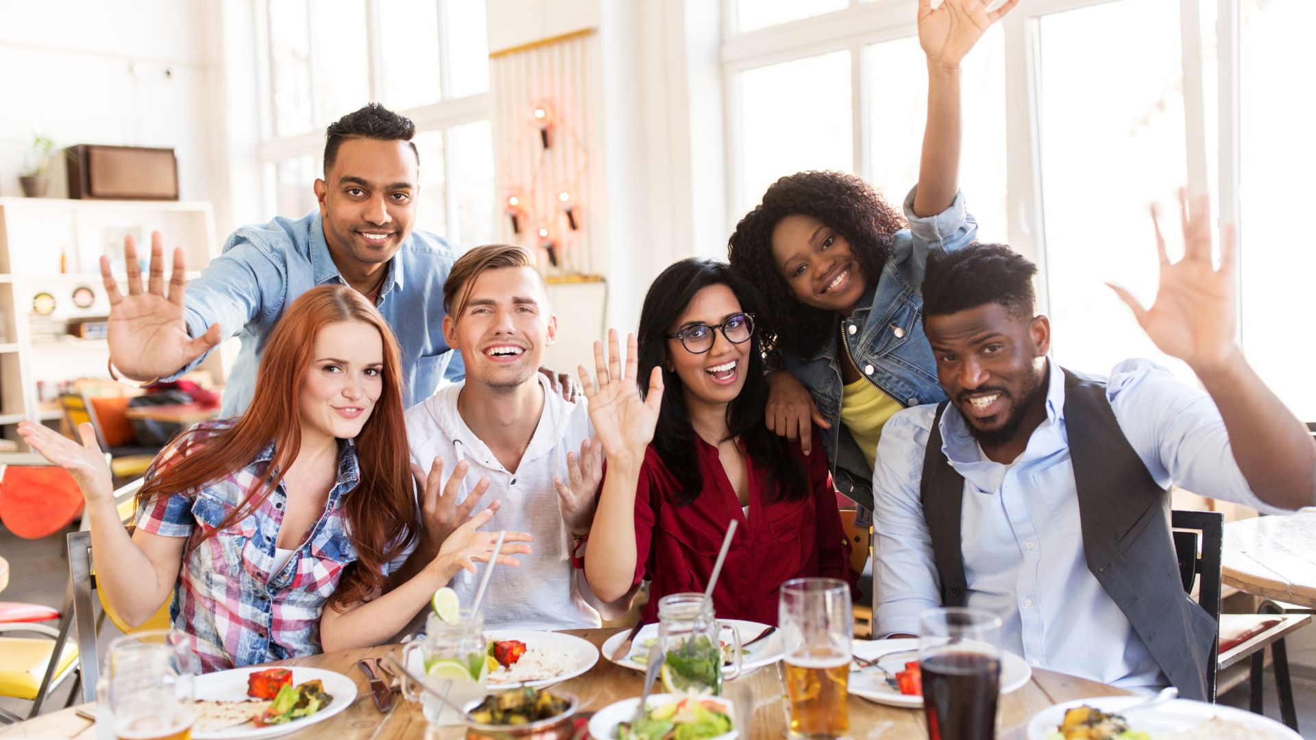 A diverse group of friends waving and celebrating together at a restaurant table.