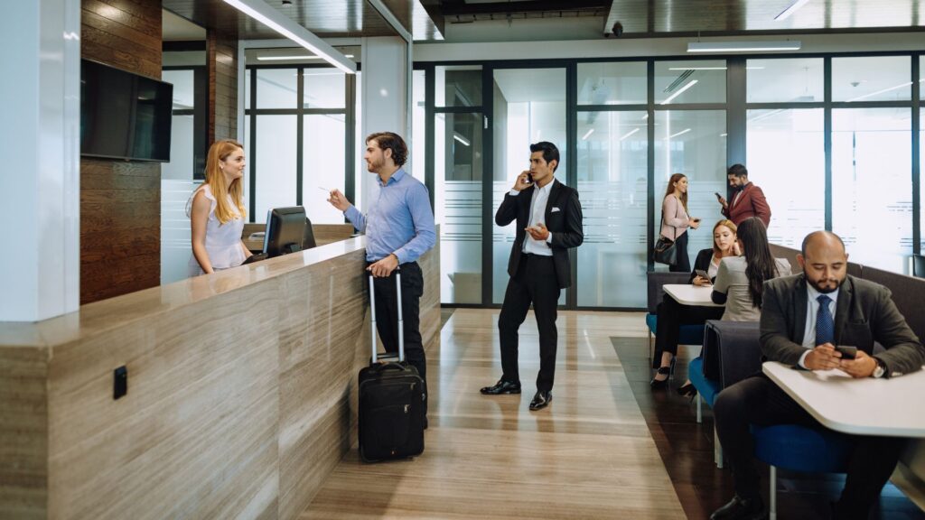 Business travelers checking in at a hotel front desk, rolling suitcase in hand — travel that's part of a regular routine